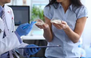 Woman considering breast revision surgery holds breast implants with plastic surgeon during consultation